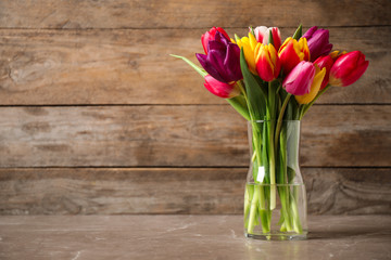 Beautiful spring tulips in vase on table against wooden background. Space for text