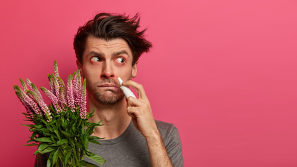 Studio shot of sick man has red watery eyes, drips nose with special medications, focused aside,...
