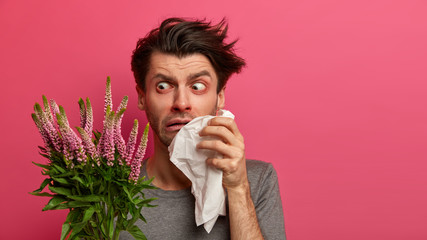 Photo of displeased allergic man reacts surprisingly at allergen, holds handkerchief near runny...