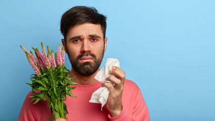 Pollen allergy symptoms concept. Puzzled unshaven man has terrible allergy, holds plant and paper...