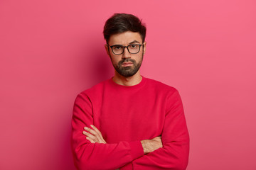 Photo of self assured man with beard, keeps arms folded over body, looks seriosly at camera, wears casual clothes, has talk with colleague, poses indoor against pink background. Confident guy