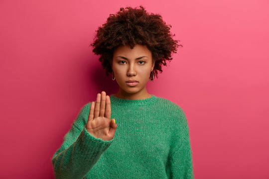 Young serious curly haired Afro American woman does stop sign with palm, wears green sweater, demonstrates prohibition and restriction, refuses something, models against pink background, says no