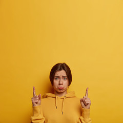 Upset depressed European woman waits for explanation, looks sadly at camera, points fingers up, demonstrates something unpleasant upwards, purses lips, wears hoodie, models over yellow studio wall