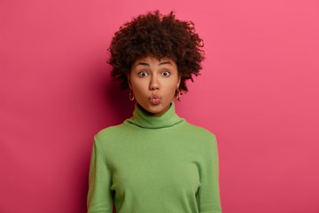 Fototapeta na wymiar Beautiful woman with natural curly hair, healthy skin, keeps lips folded, gazes directly at camera, wears neat green turtleneck, models against bright pink background, wants to kiss someone.