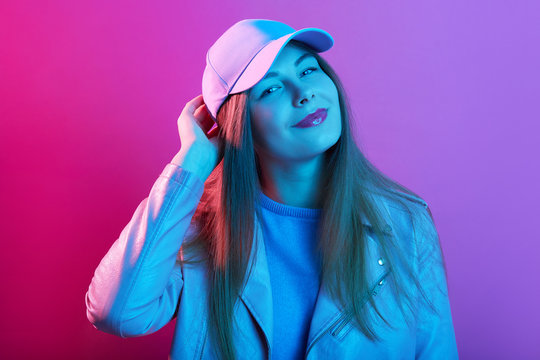 Horizontal indoor image of attractive magnetic young female standing isolated over neon background in studio, wearing cap, sweatshirt and jacket, looking directly at camera, smiling sincerely.