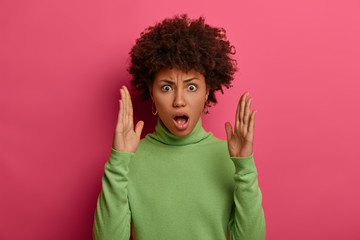 Shocked displeased curly woman raises palms, shapes very big item, tells about size, opens mouth from wonder, wears green poloneck, isolated over vibrant pink background. Body language concept