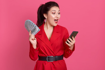 Indoor image of busy emotional charming young female opening mouth widely, holding fan of money and smartphone in both hands, looking at device screen with surprise. People and money concept.