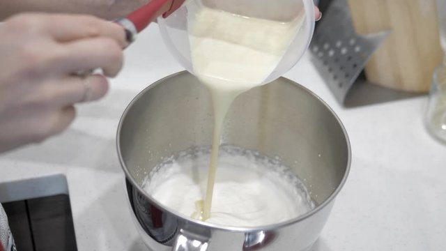 Female hands pour milk liquid into a white base in a metal cup, close-up.