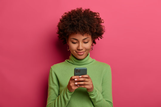 Horizontal shot of pleased dark skinned woman uses mobile phone for texting with friends, reads post online, connected to wireless internet, concentrated in display with smile, models indoor