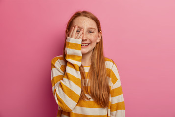 Carefree optimistic ginger girl keeps palm on face, smiles toothily, giggles as hears jokes, has long foxy hair, freckled face, wears loose jumper, isolated on pink wall. Children, emotions, laughter