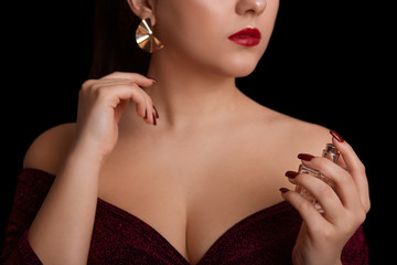 Perfume bottles in hands of beautiful girl oi studio isolated over black background, part of face of young woman with aroma, concept of care and cosmetics, female wearing elegant dress and earrings.