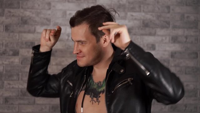 A handsome man with a tattoo on his chest in a black jacket, dancing against a gray brick wall.