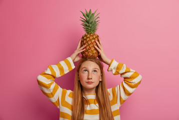 Little pretty female child holds ripe pineapple on head, has long red hair, dressed in striped jumper, poses indoor against pink background. Adorable freckled kid with tropical fruit, makes trick