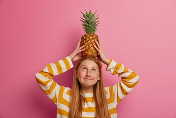 Studio shot of lovely long haired foxy girl has charming look, holds pineapple on head, concentrated above, plays with ripe fresh tropical fruit, wears white and orange striped jumper, poses indoor.