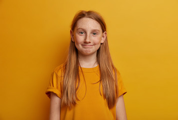 Portrait of little child presses lips, has freckled complexion, gazes straightly at camera, has positive expression, wears yellow casual t shirt, poses in studio. Children, face expressions concept