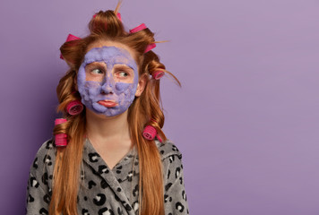 Displeased thoughtful girl focuses aside on blank space, has bad mood, wears hair curlers and...