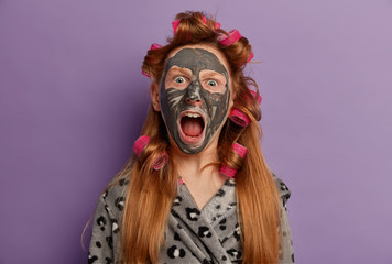 Irritated millennial girl shouts loudly, opens mouth widely, applies facial beauty mask, hair curlers, reacts on astonishing news, visits spa center, wears bathrobe, isolated on purple background