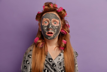 Photo of embarrassed ginger teenager gazes surprised at camera, opens mouth, expresses disbelif, wears nourishing mask on face, hair curlers, being very emotive, isolated on purple background