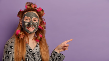 Horizontal shot of cheerful girl has toothy smile, applies clay mask for healthy complexion, wears curlers, soft robe, indicates at empty space on purple background, advertises beauty product