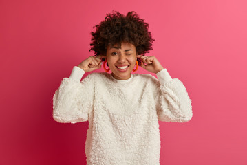 Joyful ethnic woman winks eye, smiles broadly, plugs fingers into ear holes, ignores loud noise, clenches teeth, wears winter sweater, models against pink background. Dont want listen anymore