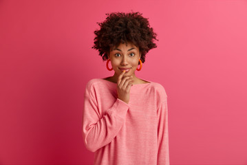 Fototapeta na wymiar Portrait of pleasant looking cheerful Afro American woman touches lips, looks curiously at camera, glad to hear good news, wears casual pink jumper, models indoor. Human expressions, emotions.