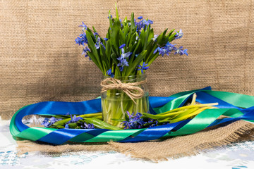 On a jute cloth on a metal tray is a jar with a bouquet of snowdrops. The composition is framed by a green and blue ribbon.