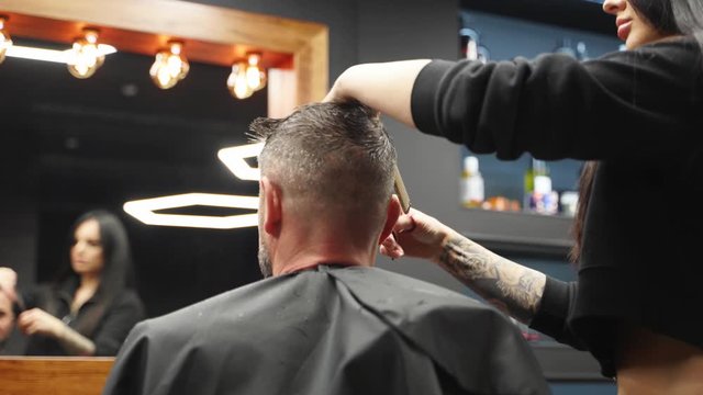Attractive barber girl cutting man's hair with scissors and comb. Female hair cutter working serving client. Stylish man sitting in vintage barber shop chair. Hairstylist woman doing trendy haircut.