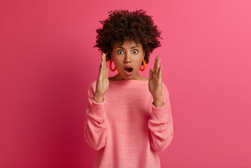 Amazed pleasant looking African American woman shocked by very big size of item, explains what shape of box she seeks, opens mouth from wonder, wears rosy jumper, isolated on pink background