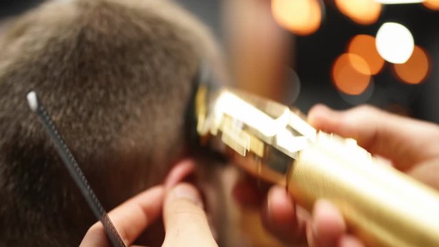 Trendy barber cuts bearded man's hair with a clipper in barbershop. Men's hairstyling and hair cutting in salon. Grooming the hair with trimmer. Hairdresser doing haircut in retro hair salon.
