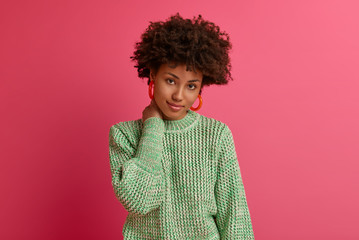 Portrait of calm curly woman touches neck, looks directly at camera with serious expression, listens employee carefully, wears knitted sweater, poses against rosy vibrant wall, has natural beauty
