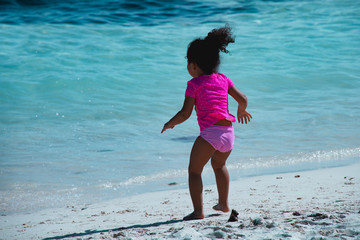 Sweet caribbean girl playing on the beach. Contrast with the intense blue color of the sea and the bright pink bathing suit and tee shirt of the girl. Vacation mood.