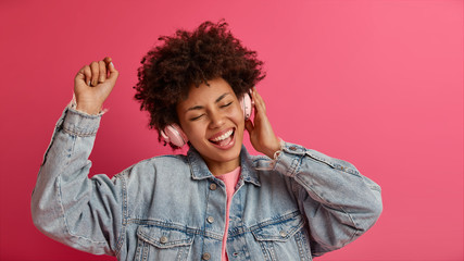 Carefree adorable woman raises arm, listens awesome new song in headphones, keeps eyes closed, moves with rhythm, wears denim jacket, isolated on pink wall. Music, lifestyle, people concept.
