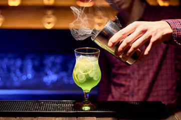 The bartender releases smoke from a shaker into a glass with a green cocktail.