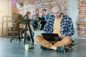Hip worker in a startup coding with laptop sitting on skateboard