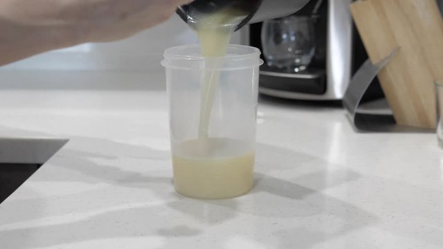 Female hands pour milk white liquid into a plastic cylinder, on a white table, closeup.