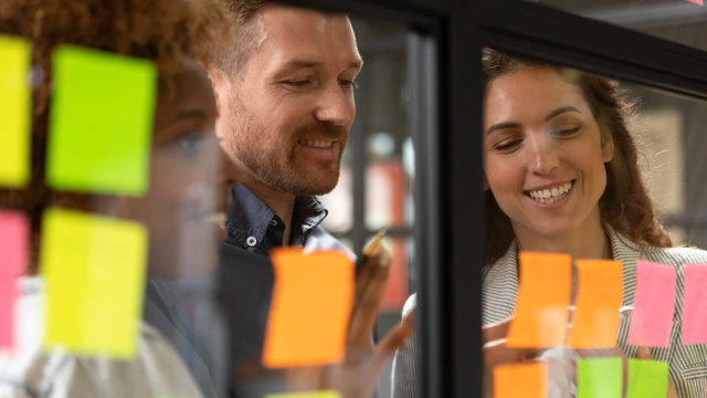 Creative diverse employees team writing ideas on sticky papers on glass wall close up, multiethnic colleagues brainstorming, working on new project plan together at briefing, looking at stickers