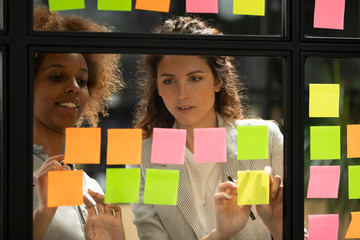 Diverse colleagues discussing notes on sticky papers close up, managers planning new project together, African American businesswoman writing creative ideas, tasks on sticker on glass scrum board