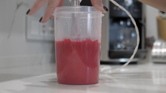 Female hands mix pink liquid with red paint, plastic cylinder, blender, closeup.