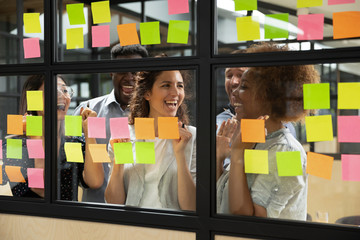 Excited colleagues celebrating success, business achievement, team victory, standing behind glass wall with colorful stickers at scrum meeting, happy colleagues finished corporate project