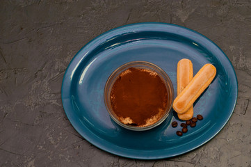Dessert of italian cuisine tiramisu with cocoa and savoyardi cookies in glass bowl on a blue plate, gray concrete background, top view, copy space