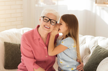 Cute girl whispering something to her grandmother's ear in living room