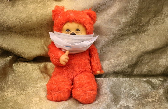 A red Teddy bear looking up in a medical mask sits on a green cloth and holds a fist with a raised thumb.