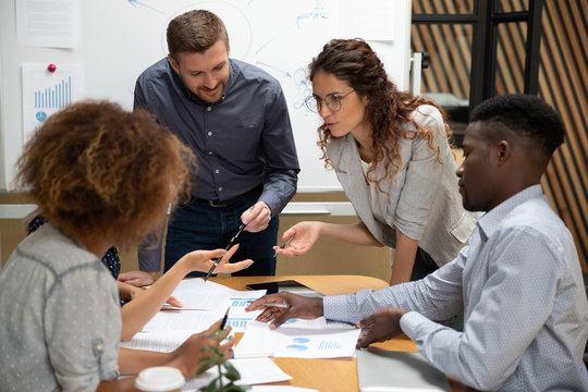Diverse employees team with leader discussing project strategy or results, statistics close up, reading documents at meeting in boardroom, business partners sharing startup ideas at briefing