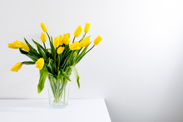 bouquet of yellow tulips is on the table opposite white wall.