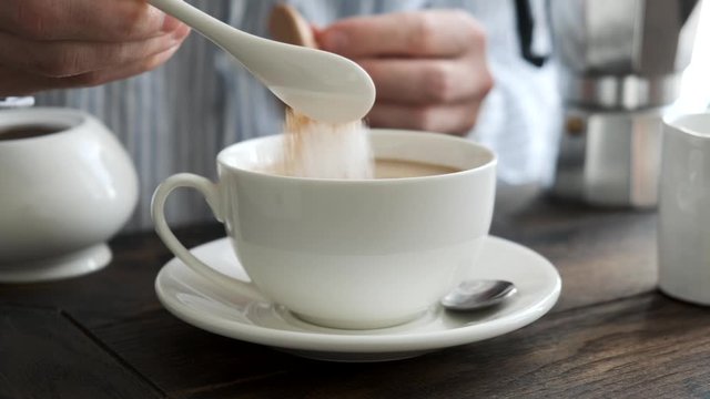 Person adding sugar into cup of coffee with milk. Wooden table background. Morning cup of coffee