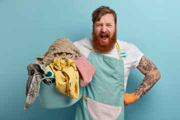 Photo of annoyed bearded man busy with housework, holds basket full of laundry and detergent, wears...