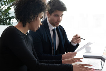 Concentrated multiracial colleagues brainstorm cooperate discuss company business project working on laptop, focused diverse businesspeople consider paperwork finances, using computer gadget together
