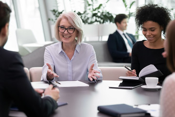 Smiling diverse multiethnic businesspeople sit at desk in office brainstorm consider financial paperwork together, happy multiracial colleagues talk discuss project at meeting, collaboration concept