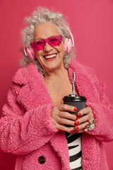 Joyful of fashionabele senior woman wears pink sunglasses and fur coat, has red manicure and makeup, drinks takeaway coffee, listens music in headphones. Old people, modern technologies concept