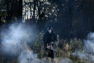 Glaring evil man in black coat with a chainsaw in a misty forest in the Fall at Halloween